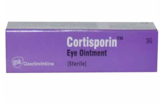 A tube of Cortisporin Eye Ointment placed on a white surface with scattered eye-related medical equipment in the background.